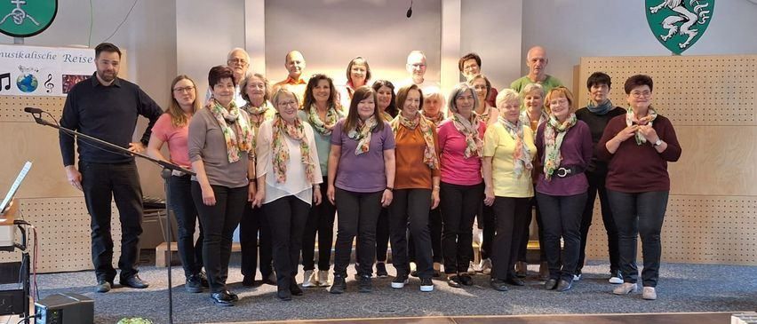 Ein Chor von Erwachsenen, sowohl Männer als auch Frauen, posiert für ein Foto auf der Bühne mit Schals um den Hals. Ein Mikrofon und ein Lautsprecher sind auf der linken Seite aufgestellt.