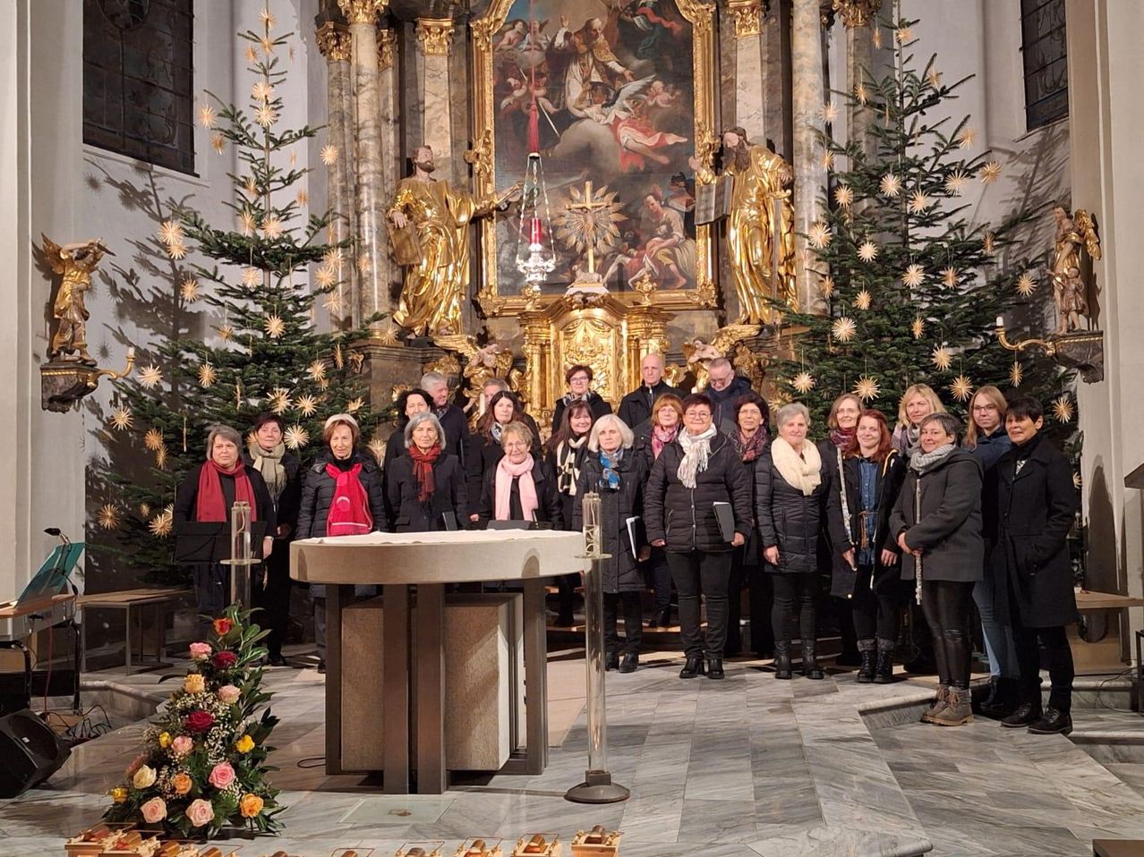 Ein Chor steht vor einem Kirchenaltar, geschmückt mit Weihnachtsbäumen und Blumen, mit einem goldenen Gemälde dahinter.