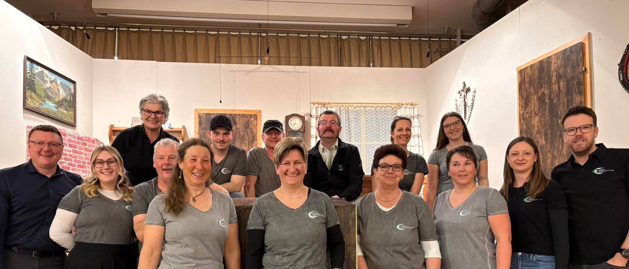 Eine Gruppe von Menschen in passenden grauen T-Shirts posiert für ein Foto in einem Kunststudio mit Holzwänden und einer Uhr an der Wand.