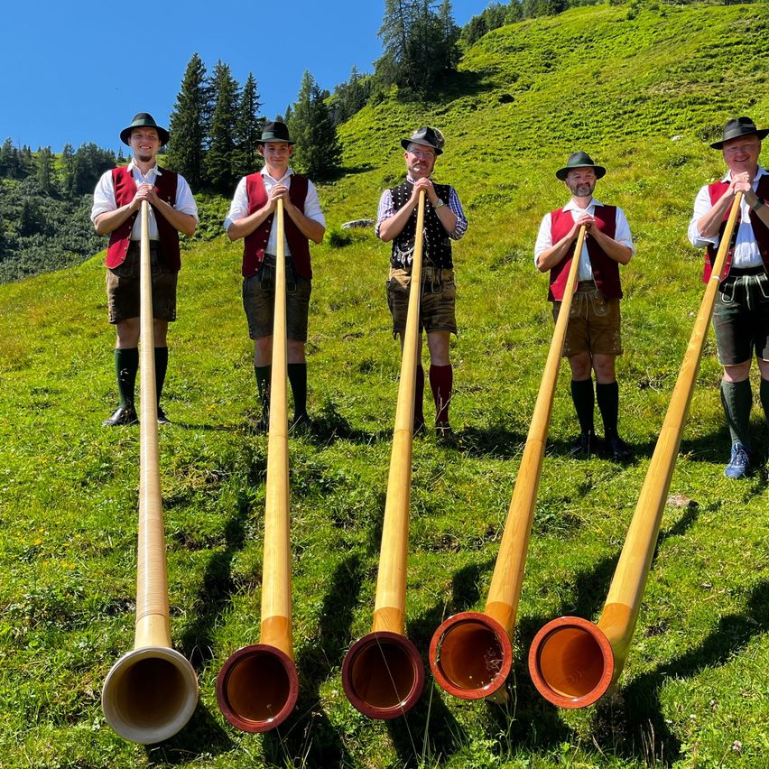 Fünf Männer in traditioneller Kleidung stehen auf einem Grashügel und halten lange Holzhörner. Sie nehmen wahrscheinlich an einer kulturellen Veranstaltung teil.
