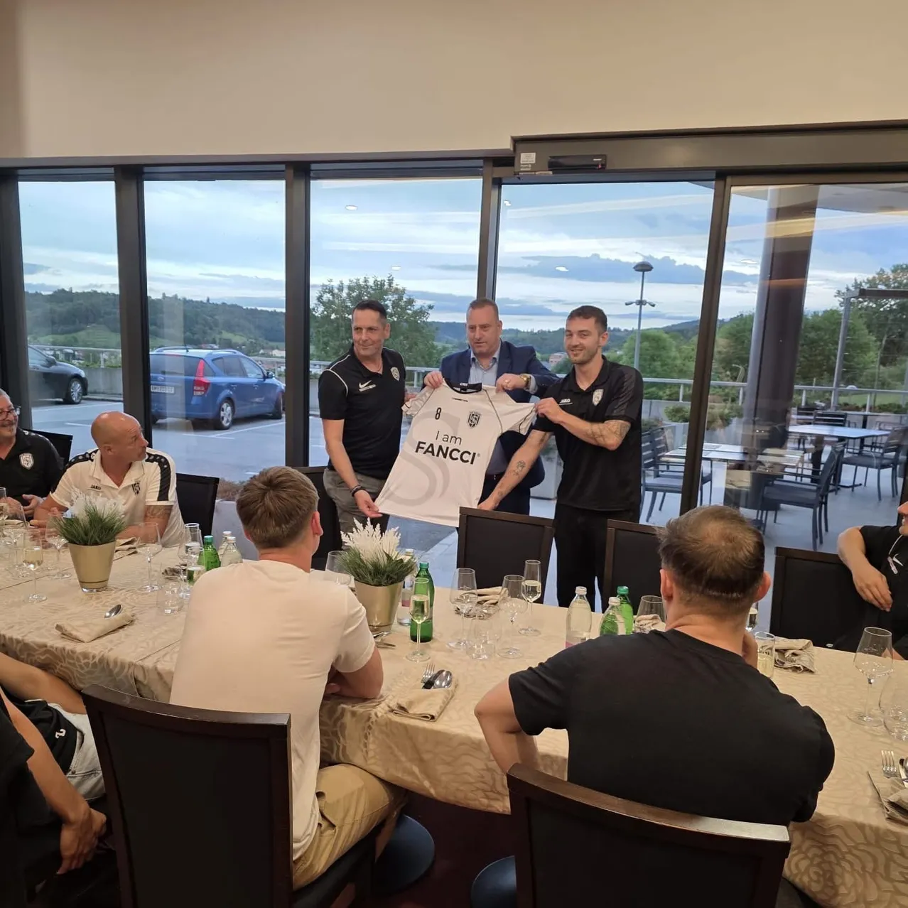 Eine Gruppe von Männern versammelt sich um einen Tisch mit einem weißen Shirt, auf dem 'Ich bin Fanci' steht. Sie lächeln und scheinen zu feiern. Der Tisch ist mit Weingläsern, Flaschen und Topfpflanzen gefüllt. Hinter ihnen befinden sich Stühle und Tische mit sitzenden Personen. Draußen am Fenster sind Autos und Bäume zu sehen.