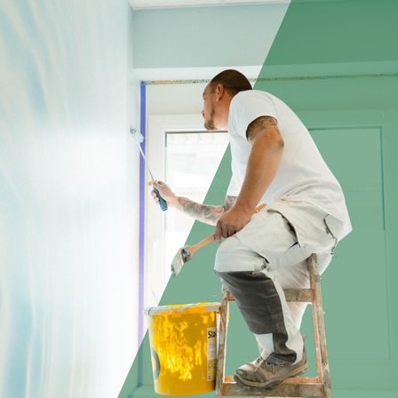 Ein Mann malt eine Wand und benutzt einen Pinsel, ein gelber Farbkübel steht vor ihm.