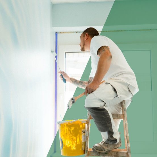 Ein Mann malt eine Wand und benutzt einen Pinsel, ein gelber Farbkübel steht vor ihm.