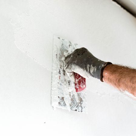 Eine Person in Handschuhen trägt Putz mit einem Spachtel auf eine weiße Wand auf. Der Putz ist weiß mit einer roten Linie. Die Wand ist glatt und sauber.