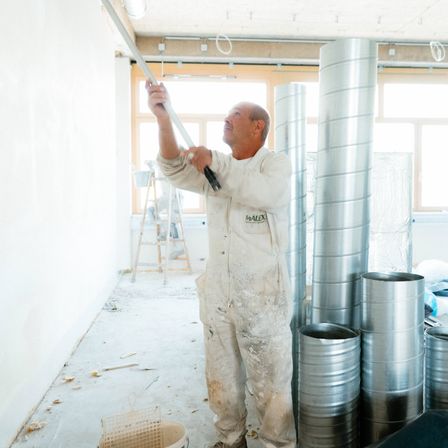 Ein Mann in weißen Overalls malt eine Wand mit einem Wischer, ein Eimer in der Nähe. Rohre und Fenster sind im Hintergrund zu sehen.