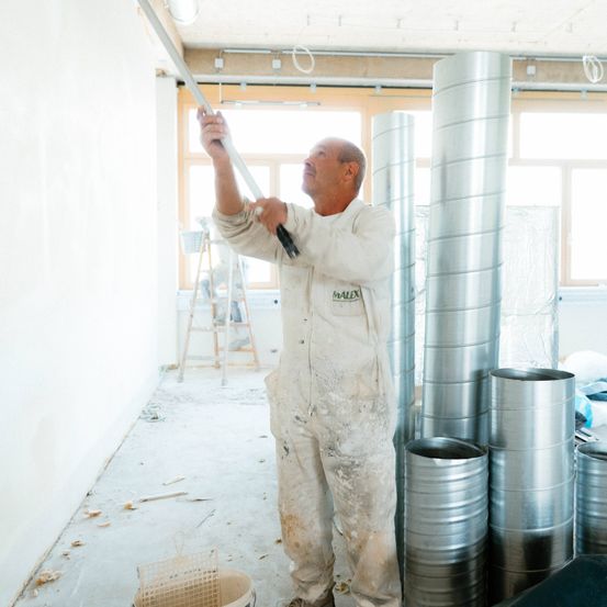 Ein Mann in weißen Overalls malt eine Wand mit einem Wischer, ein Eimer in der Nähe. Rohre und Fenster sind im Hintergrund zu sehen.