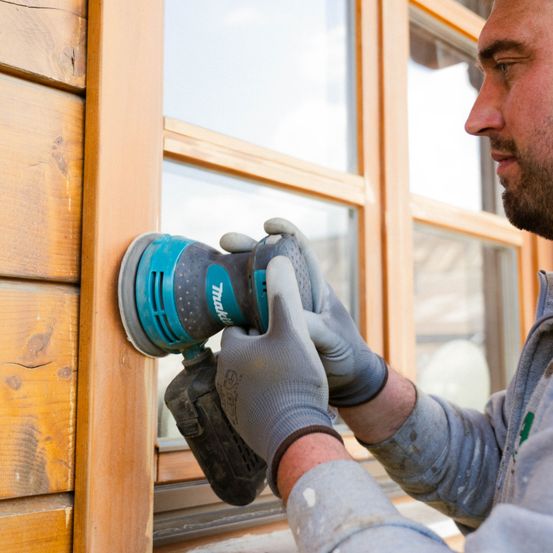 Ein Mann in grauer Arbeitskleidung und Handschuhen schleift eine Holzoberfläche mit einem elektrischen Schleifer.