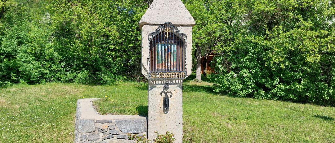 Ein Betonpfeiler mit einem Metalltor und einem Kreuz oben steht auf einer Grasfläche.