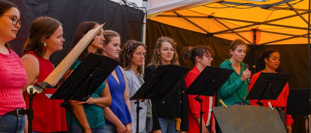 Ein Mädchenchor übt in einem Zelt mit Notenständern und Instrumenten. Sie sind in verschiedenen Farben gekleidet und scheinen sich auf eine Aufführung vorzubereiten.