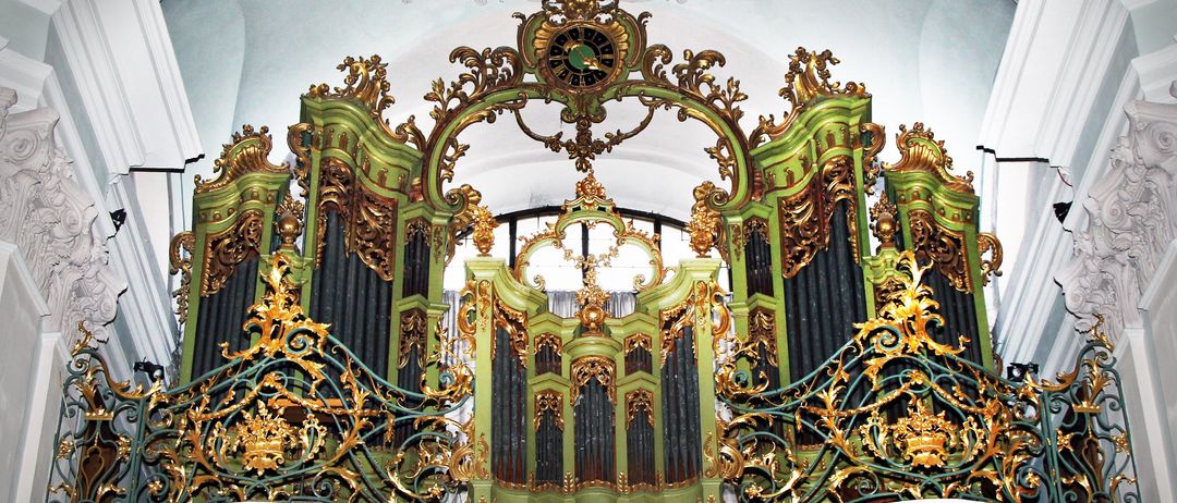 Eine kunstvolle grüne und goldene Orgel hängt von der Decke einer Kirche. Die Decke hat ein Gemälde eines Engels.