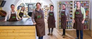 Fünf Frauen in braunen Schürzen stehen lächelnd in einem Geschäft. Hinter ihnen sind Regale mit Produkten zu sehen. Im Vordergrund befindet sich eine Holztheke.