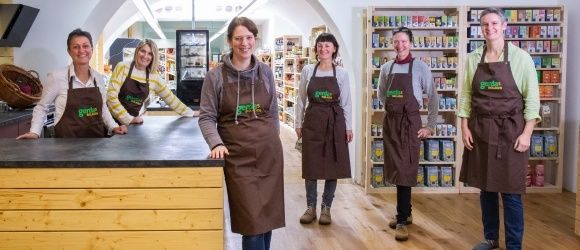 Fünf Frauen in braunen Schürzen stehen lächelnd in einem Geschäft. Hinter ihnen sind Regale mit Produkten zu sehen. Im Vordergrund befindet sich eine Holztheke.