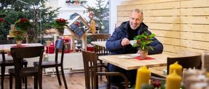 Ein Mann sitzt an einem Tisch in einem weihnachtlich dekorierten Café, hält eine Tasse und lächelt. Auf dem Tisch stehen eine Pflanze und Kerzen. Im Hintergrund sind Holzwände und Weihnachtsbäume zu sehen.
