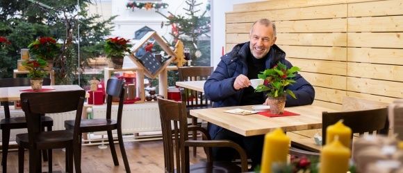 Ein Mann sitzt an einem Tisch in einem weihnachtlich dekorierten Café, hält eine Tasse und lächelt. Auf dem Tisch stehen eine Pflanze und Kerzen. Im Hintergrund sind Holzwände und Weihnachtsbäume zu sehen.