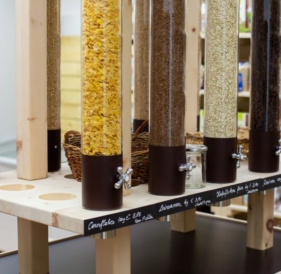 Ein hölzernes Regal zeigt verschiedene Getreidesorten in transparenten Behältern, die mit ihren Namen und Preisen beschriftet sind.