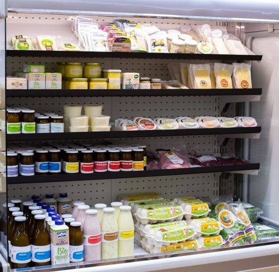 Ein Kühlregal in einem Geschäft zeigt eine Vielzahl von Produkten, darunter Milchprodukte, Fleisch und Gemüse. Die Regale sind mit Gläsern, Flaschen und Eiern bestückt.