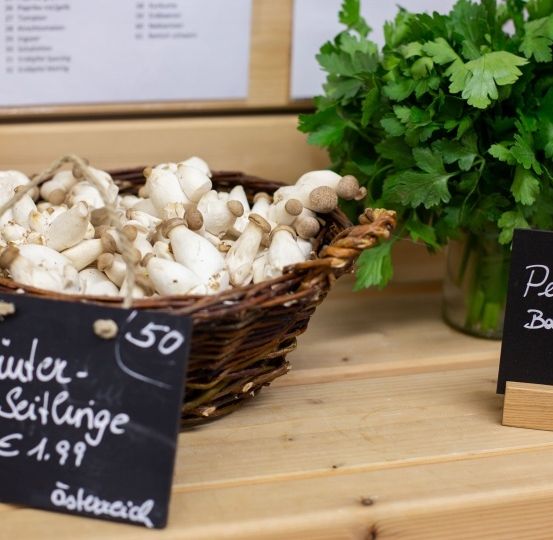 Auf einem Holztisch stehen ein Korb mit weißen Pilzen und eine Vase mit Petersilie. Ein Schild bietet Pilze für 1,99 Euro pro 100 Gramm an.