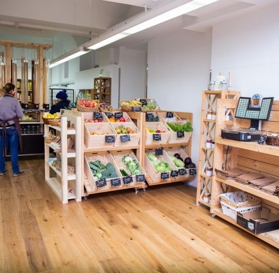 Ein Blick ins Innere eines Lebensmittelgeschäfts mit Holzregalen voller Gemüse und Obst, eine Person mit Maske steht da.
