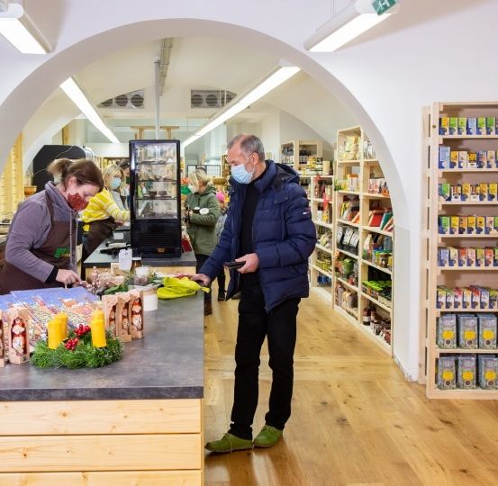 In einem Laden steht ein Mann mit Maske neben einem Tresen mit Lebensmitteln, während eine Frau hinter dem Tresen steht. An den Wänden stehen Regale mit Waren gefüllt.