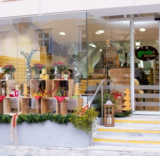 Das Geschäft zeigt ein festliches Weihnachtsthema mit Holzregalen, Weihnachtsbäumen und Pflanzen. Der Innenraum ist durch die Glastüren sichtbar, und Regale mit Produkten befinden sich im Inneren.