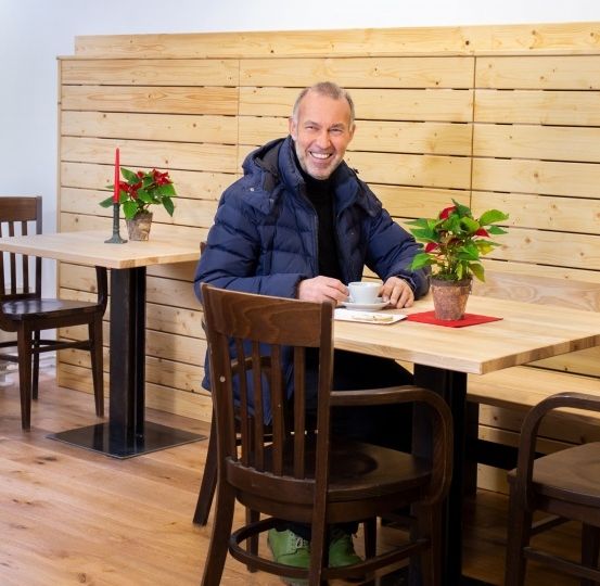 Ein Mann in einer blauen Jacke sitzt an einem Tisch mit einer Tasse Kaffee und lächelt. Blumentöpfe und eine Kerze schmücken den Tisch. Hinter ihm befindet sich eine Holzwand mit einem sichtbaren Weihnachtsbaum.