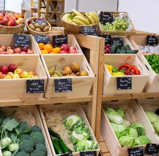 Ein Holzregal mit verschiedenen Kisten von Obst und Gemüse. Die Kisten sind mit Preisen beschriftet, darunter Äpfel, Bananen, Orangen und Trauben. Es gibt auch Kisten mit Birnen, Tomaten und Brokkoli.
