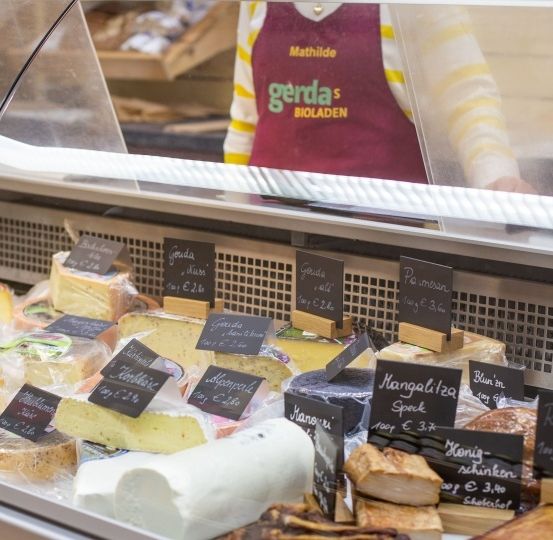 Eine Vitrine mit verschiedenen Käsesorten, einige mit Preisschildern und einer Person im Hintergrund, die eine rote Schürze trägt.