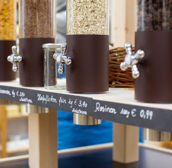Eine Theke mit mehreren braunen Spendern, gefüllt mit Körnern und Samen. Jeder Spender hat einen silbernen Hebel und die Namen und Preise der Produkte sind auf einem Schieferbrett handgeschrieben.