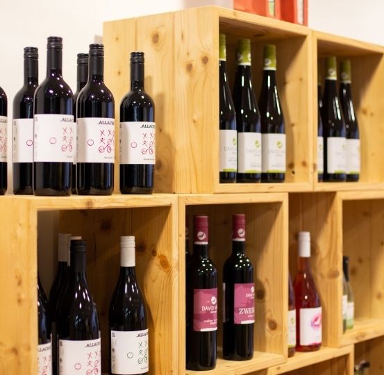 Ein Holzregal zeigt mehrere Weinflaschen, einschließlich Rot- und Weißweine, wobei einige Flaschen weiße Etiketten mit Text und Logos haben.