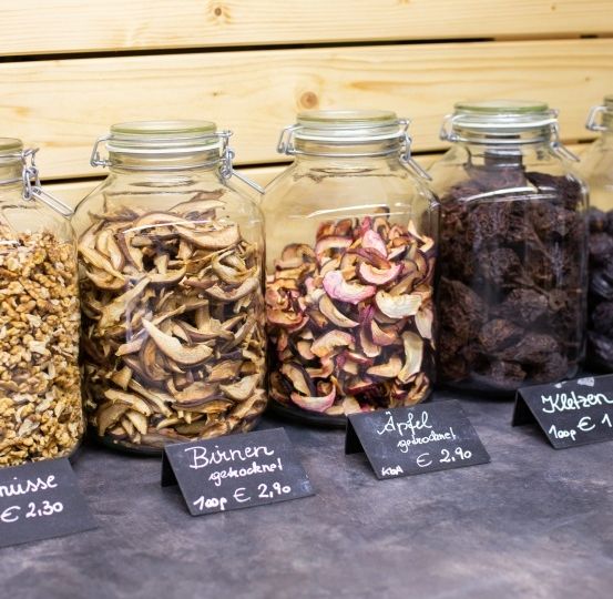Fünf Glasgefäße mit verschiedenen getrockneten Früchten und Nüssen stehen auf einer Holzoberfläche. Von links nach rechts enthalten die Gläser Walnüsse, Pilze, Äpfel, Rosinen und Pflaumen. Jedes Gefäß hat einen Preisschild, das den Preis pro 100 Gramm angibt.