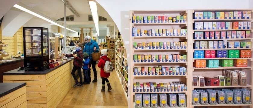 Eine Familie mit Kindern besucht einen Laden, stöbert bunte Produkte auf Holzregalen und betrachtet eine Theke und eine Glasvitrine im Hintergrund.