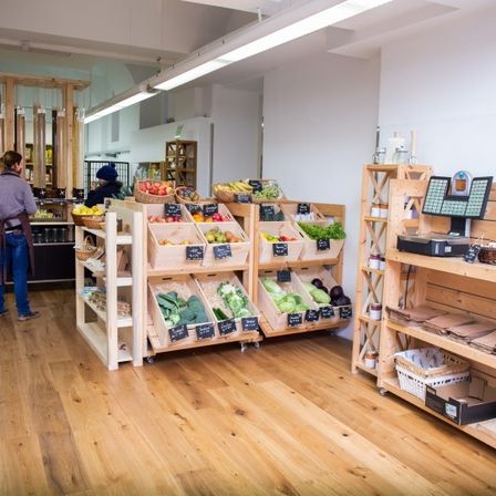 Ein Innenmarkt mit Holzregalen, die mit frischem Obst und Gemüse bestückt sind. Einige Leute sind drinnen, einige tragen Gesichtsmasken.
