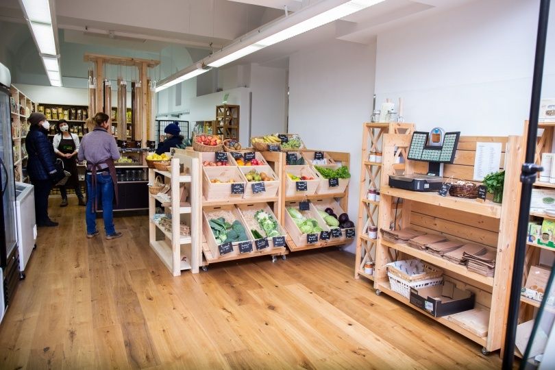 Ein Innenmarkt mit Holzregalen, die mit frischem Obst und Gemüse bestückt sind. Einige Leute sind drinnen, einige tragen Gesichtsmasken.