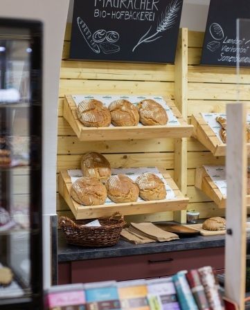 Ein Bäckereiladen mit Brot auf Regalen, einem Korb und einem schwarzen Tafelschild mit der Aufschrift Mauracher Bio-Hofbäckerei.