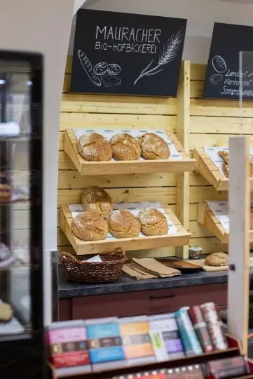 Ein Bäckereiladen mit Brot auf Regalen, einem Korb und einem schwarzen Tafelschild mit der Aufschrift Mauracher Bio-Hofbäckerei.