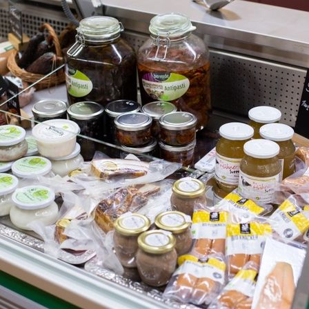 Ein gekühltes Display mit verschiedenen Lebensmitteln. Gläser mit eingelegtem Gemüse, Würsten und anderen Artikeln sind in Reihen angeordnet. Ein Schild zeigt den Preis der Würste an.