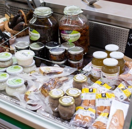 Ein gekühltes Display mit verschiedenen Lebensmitteln. Gläser mit eingelegtem Gemüse, Würsten und anderen Artikeln sind in Reihen angeordnet. Ein Schild zeigt den Preis der Würste an.