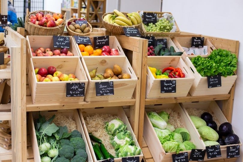 Ein Holzregal zeigt verschiedene Obst- und Gemüsesorten. Kisten halten Äpfel, Birnen, Bananen, Trauben, Tomaten, Gurken und Salat. Jede Kiste hat einen Preisschild, und das Regal ist ordentlich geordnet.