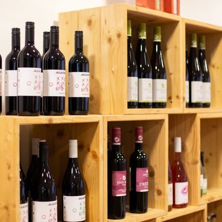 Ein hölzerne Regal präsentiert verschiedene Weinflaschen in unterschiedlichen Größen und Farben, darunter Rot, Weiß und Rosa. Die Flaschen haben weiße Etiketten mit Text und Bildern.