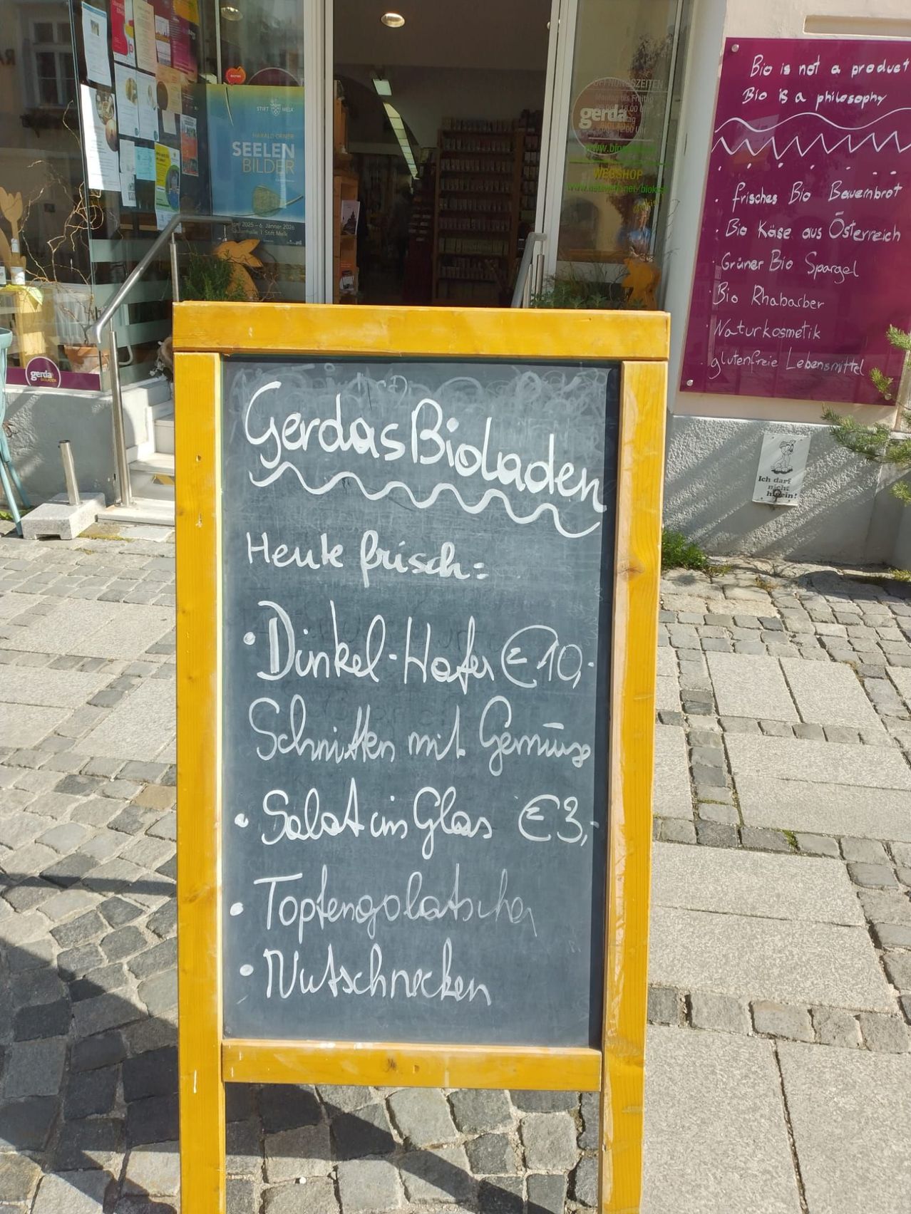 Ein gelb gerahmter Kreidetafel steht vor einem Geschäft. Sie listet die heutigen frischen Salate auf, darunter Dinkel-Hafer für 10€, Schnitten mit Germnür für 3€ und Topfengolabube. Das Geschäft hat eine Glastür und Regale, die im Inneren sichtbar sind.