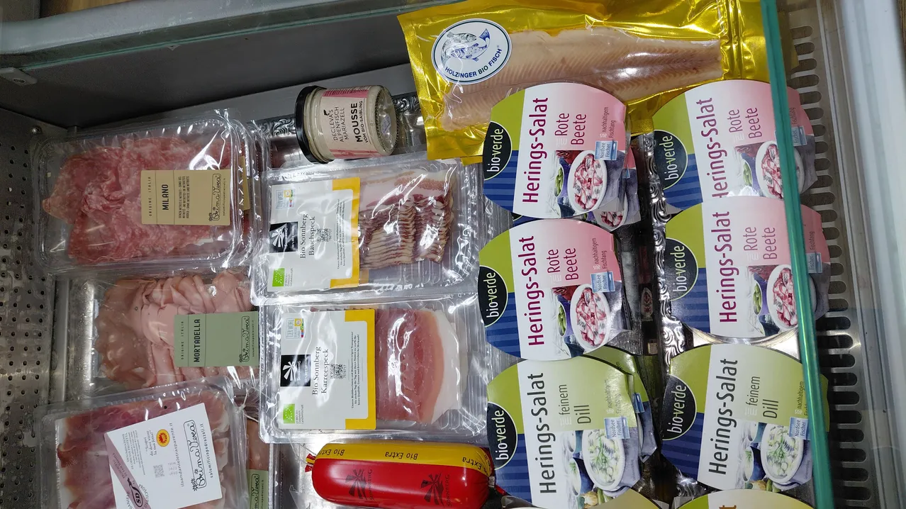 Verschiedene Packungen mit Fleisch und Fisch sind auf einem Glasregal ausgestellt, darunter Packungen von Heringssalat, Schweinefleisch und Speck. Eine Wurst wird mit dem Etikett 'Bio Salami' hervorgehoben.