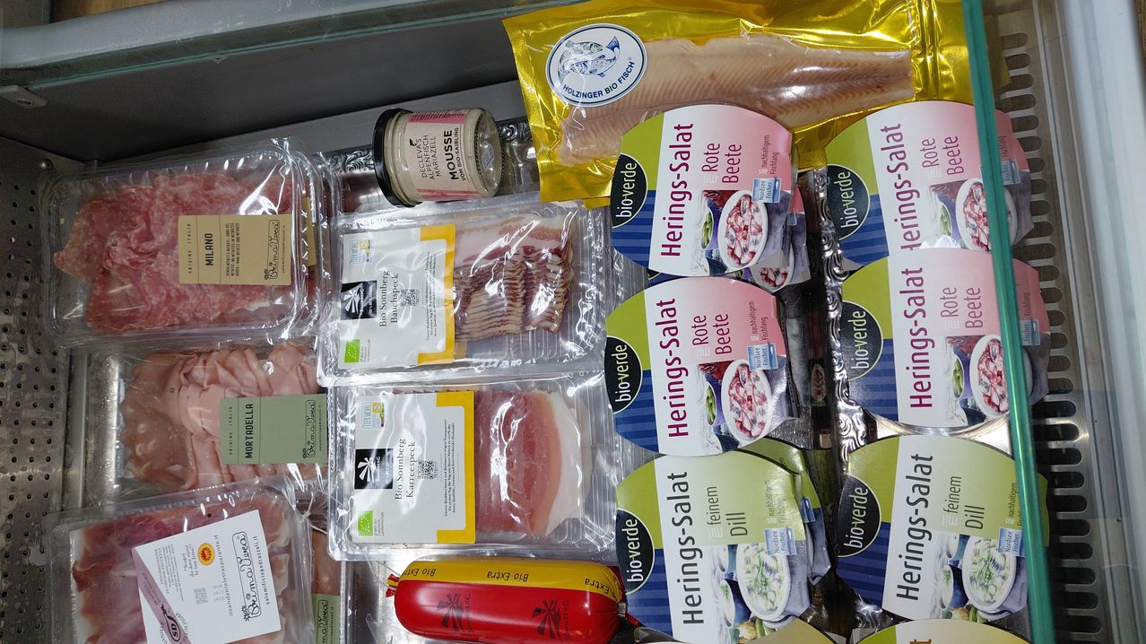 Verschiedene Packungen mit Fleisch und Fisch sind auf einem Glasregal ausgestellt, darunter Packungen von Heringssalat, Schweinefleisch und Speck. Eine Wurst wird mit dem Etikett 'Bio Salami' hervorgehoben.