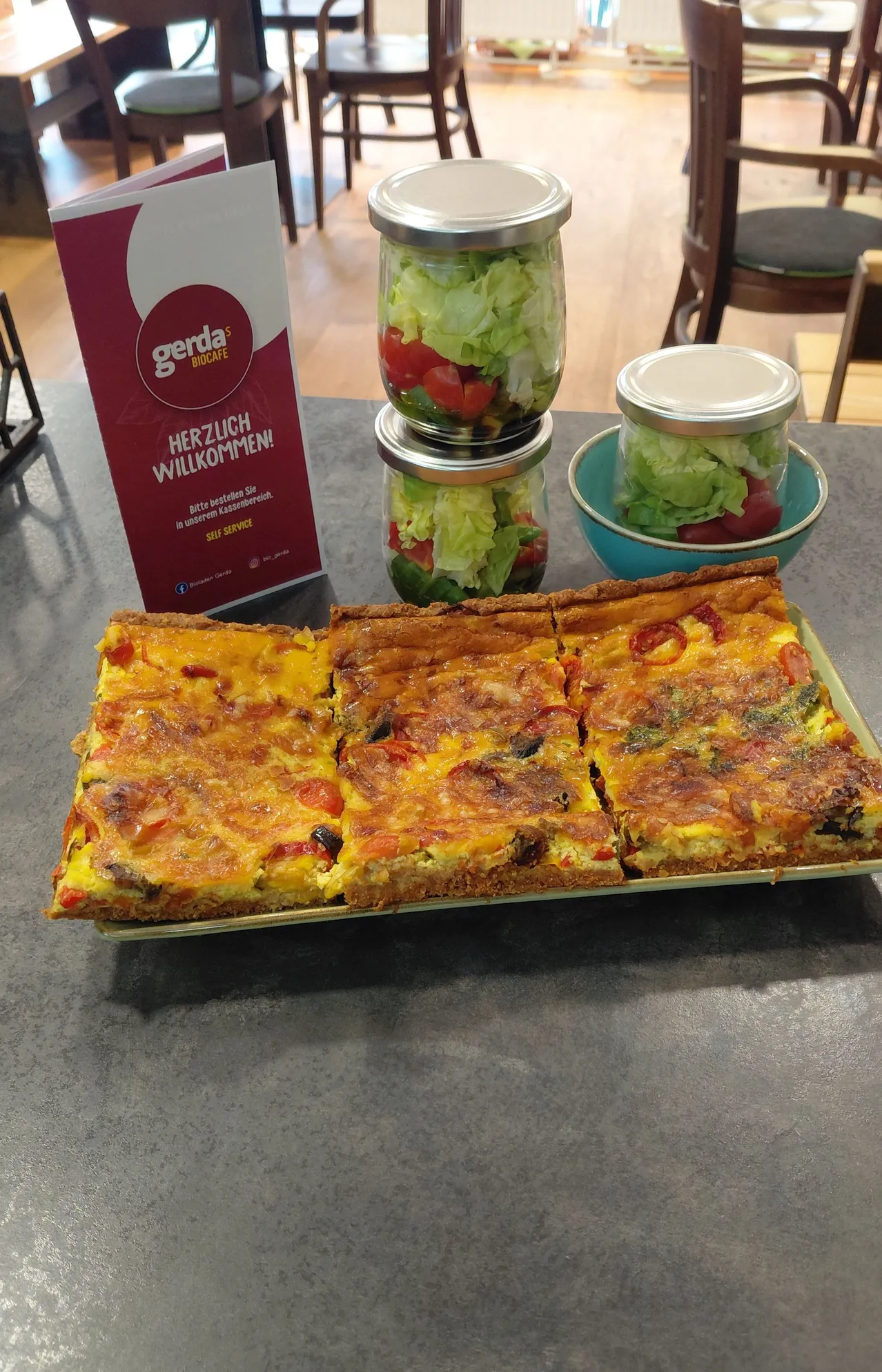 Ein Tablett mit geschnittenem Quiche mit Käse und Gemüse, begleitet von einem roten Gerda Biocafe-Schild und Salatgläsern.