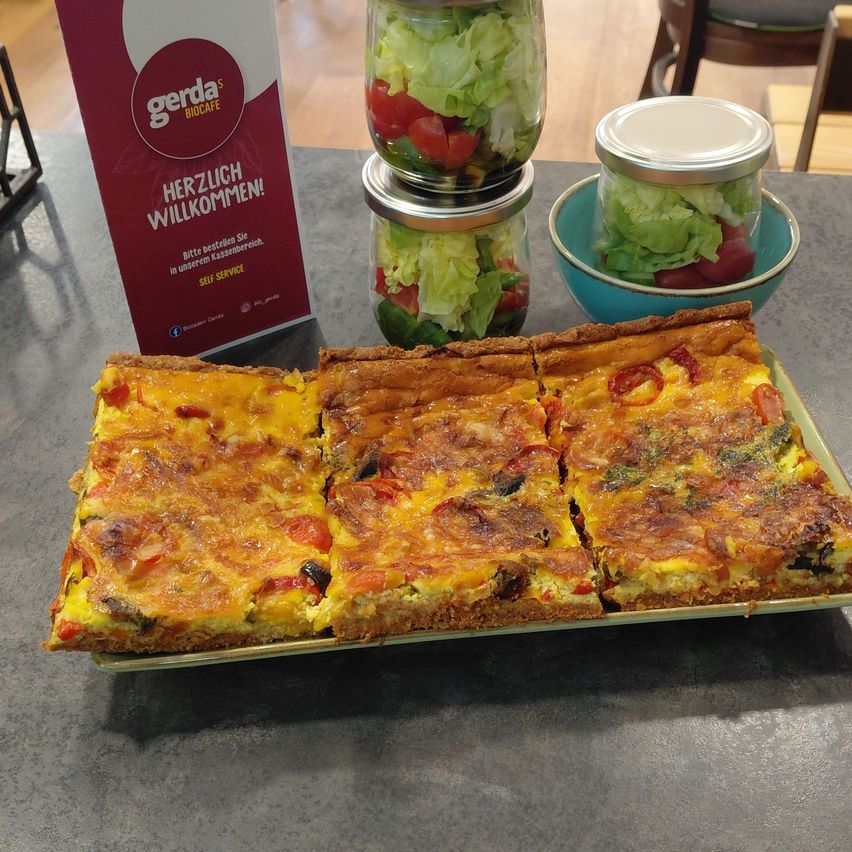 Ein Tablett mit geschnittenem Quiche mit Käse und Gemüse, begleitet von einem roten Gerda Biocafe-Schild und Salatgläsern.