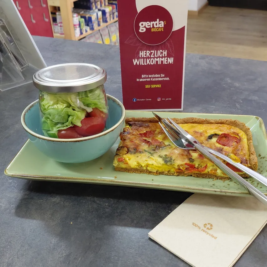 Ein Teller mit einer Quiche, einem Salat in einem Glas und einer Gabel und einem Messer auf einem Tisch mit einem Gerda's Biocafe-Schild.