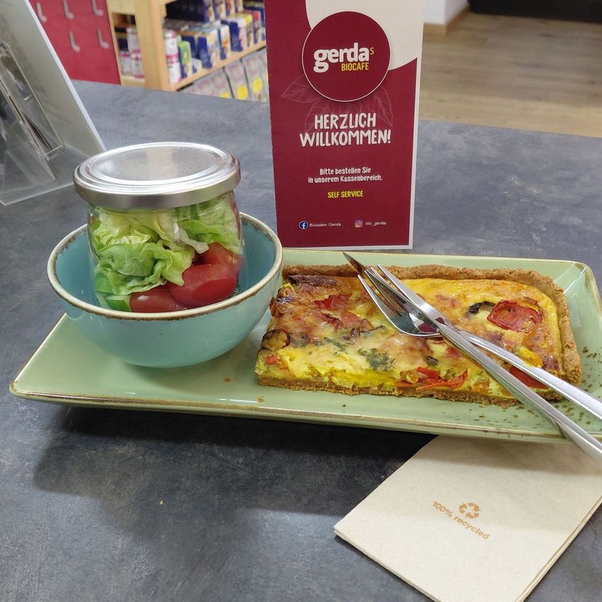 Ein Teller mit einer Quiche, einem Salat in einem Glas und einer Gabel und einem Messer auf einem Tisch mit einem Gerda's Biocafe-Schild.
