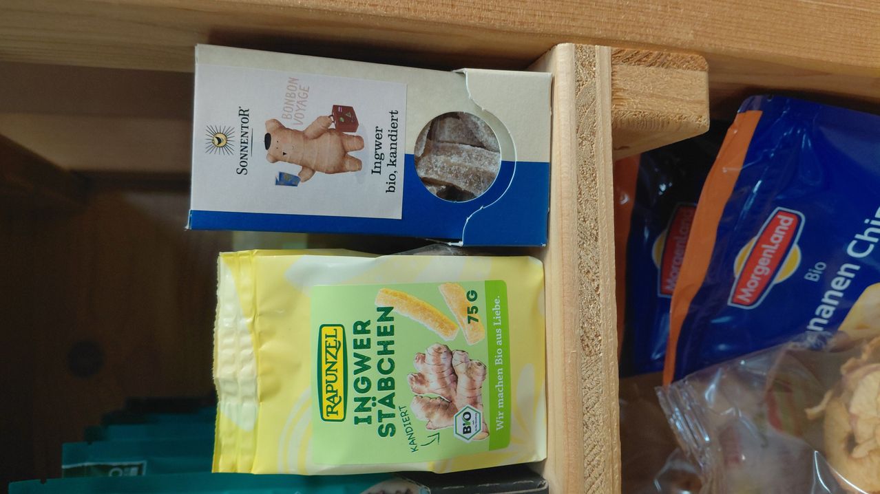 Eine Holzschublade enthält einen gelben Beutel mit Bio-Ingwer-Bonbons und eine blau-weiße Schachtel mit Bio-Süßigkeiten.
