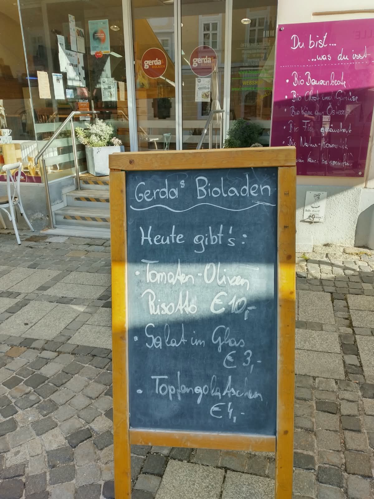 Ein Kreidetafelmenü vor einem Restaurant namens Gerda's Bioladen, das verschiedene Speisen und deren Preise auflistet.