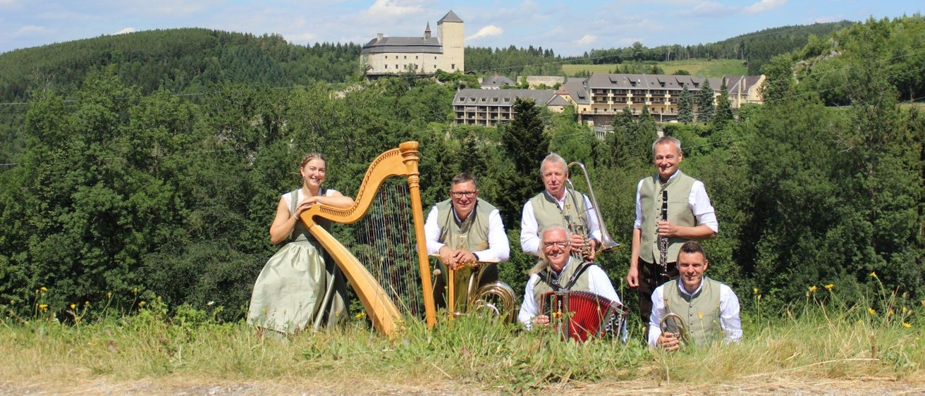 Eine Gruppe von Musikern, darunter eine Frau, die eine Harfe spielt, posiert für ein Foto vor einer malerischen Kulisse mit einem Schloss in der Ferne.