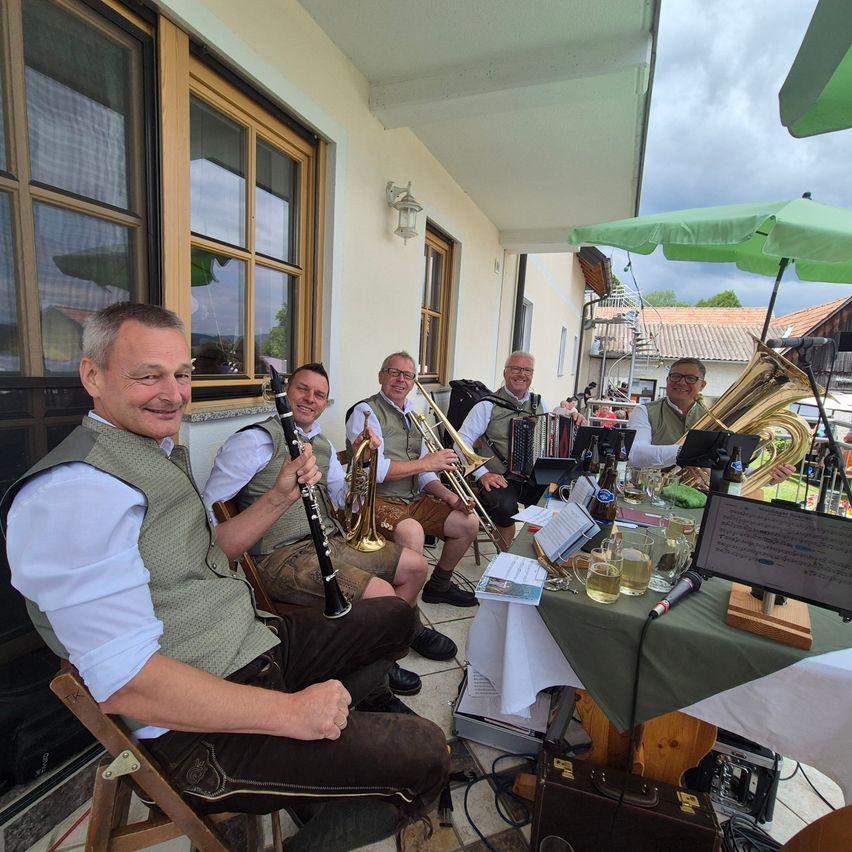 Fünf Männer in traditioneller Kleidung sitzen auf einer Veranda und spielen verschiedene Musikinstrumente. Ein Tisch mit Noten, Gläsern und einem Mikrofon steht vor ihnen.